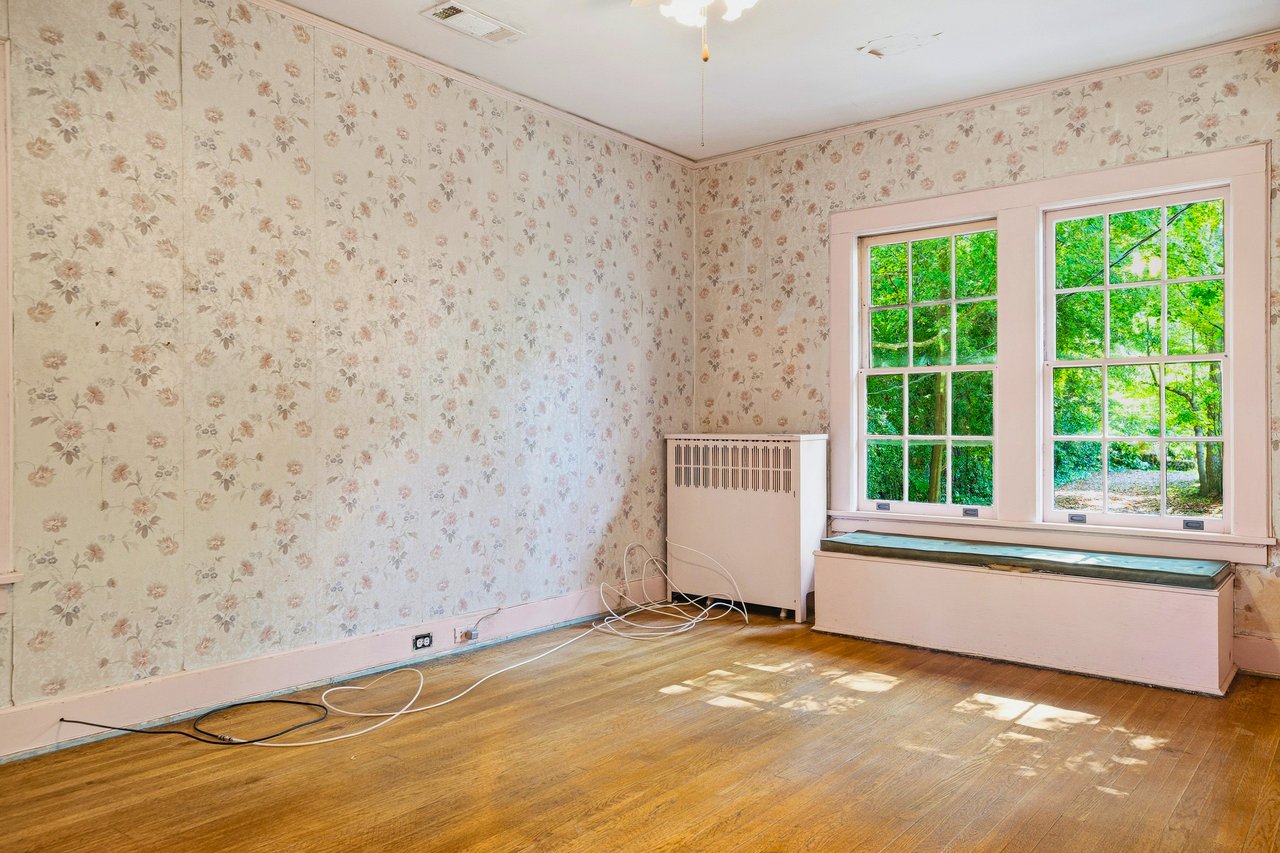 Empty room with floral wallpaper, hardwood floors, and large windows above a built-in window seat.