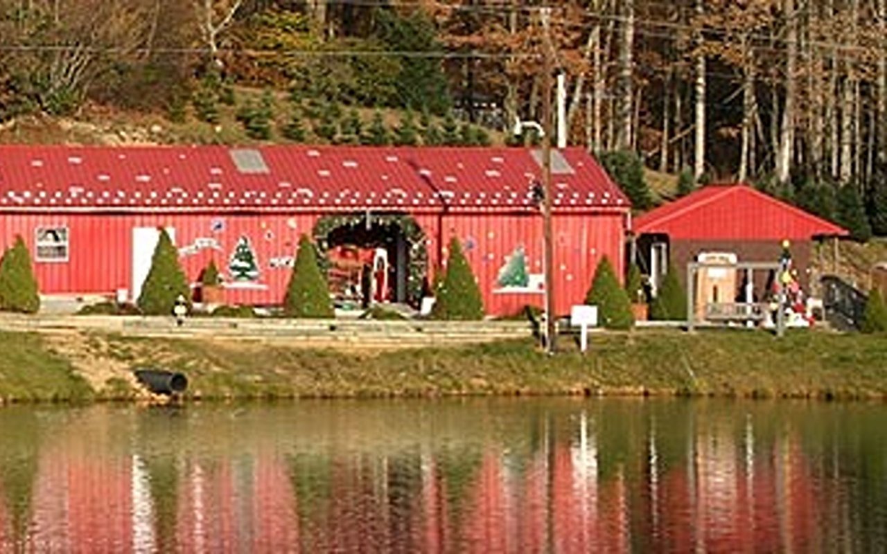Red barn decorated for Christmas beside a pond, with wooded hills behind it.