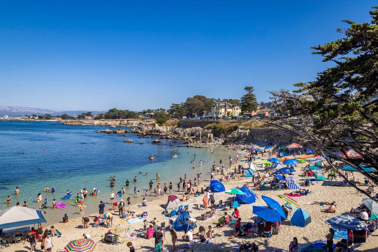 Crowded cove beach with colorful umbrellas, rocky shoreline, and coastal homes.