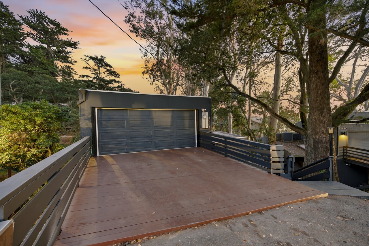 Modern dark garage and broad driveway beneath mature trees at sunset.