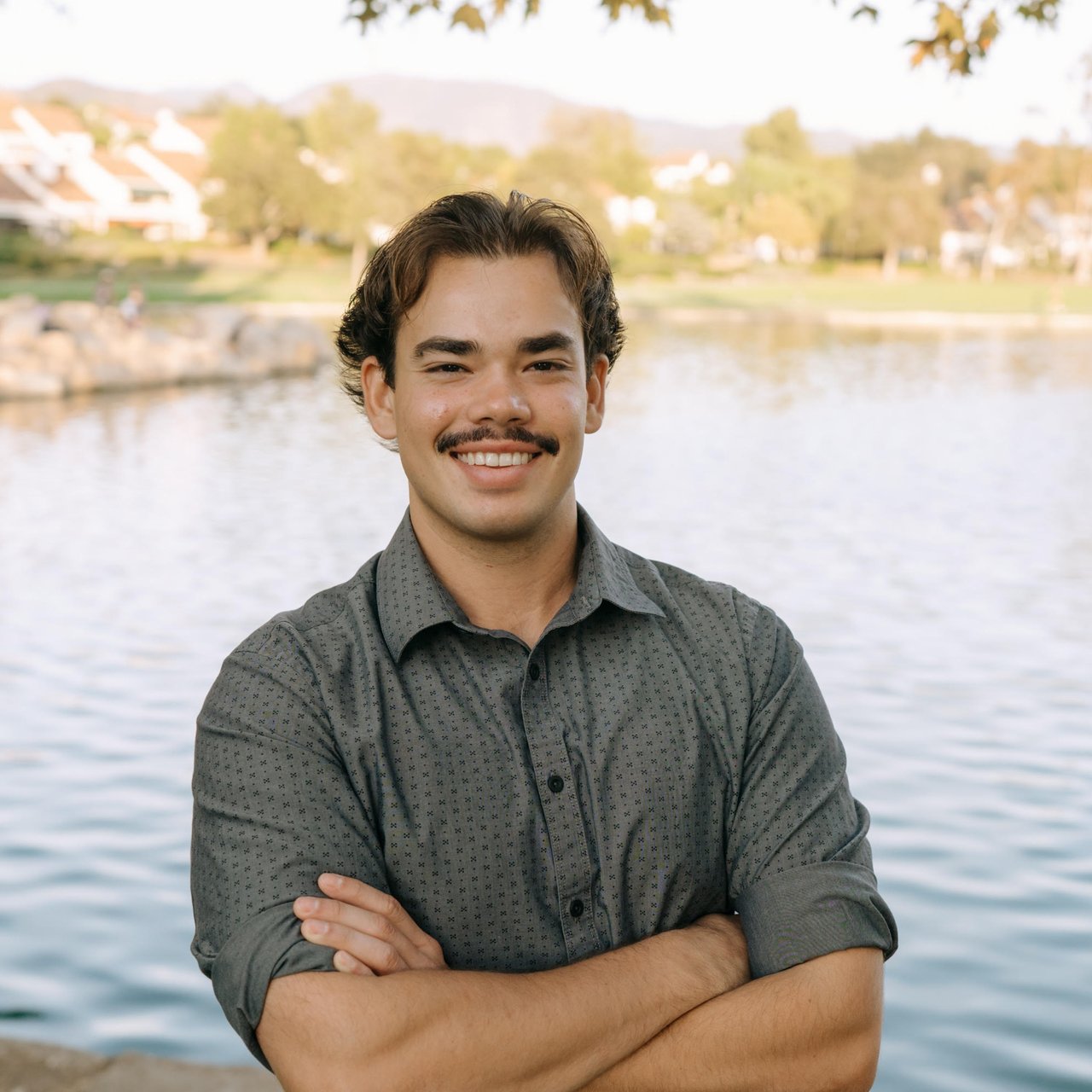 Liam Enyeart standing with crossed arms beside a pond, wearing a patterned gray shirt.