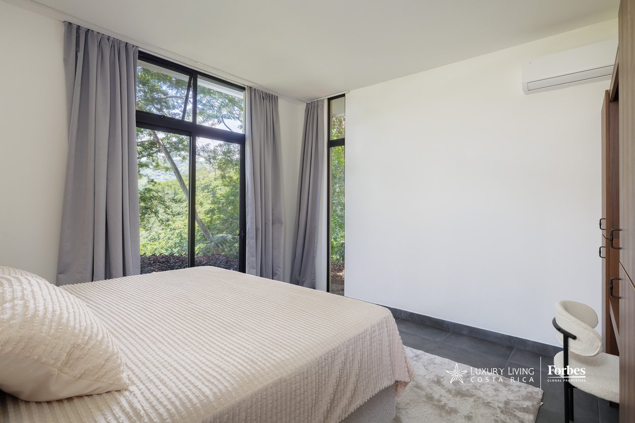 Modern bedroom with a white quilted bed and large windows overlooking greenery.