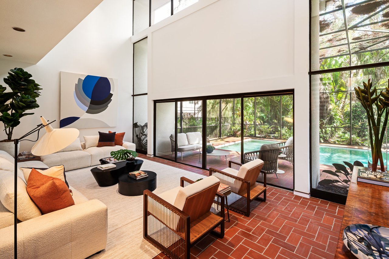 Living room with white sofas and wooden chairs overlooking a garden patio and swimming pool.