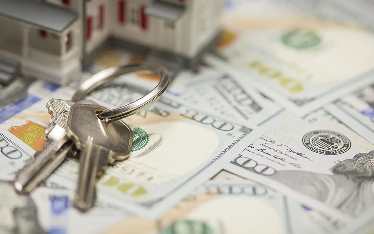 House keys resting on scattered U.S. $100 bills, with miniature houses blurred in the background.