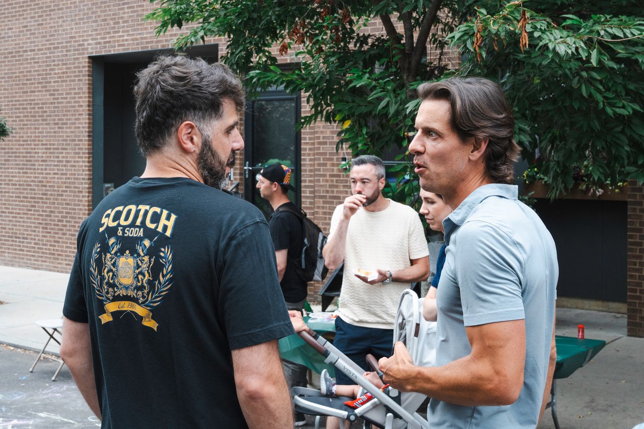 Neighbors chat at an outdoor block party beside a brick building and food tables.