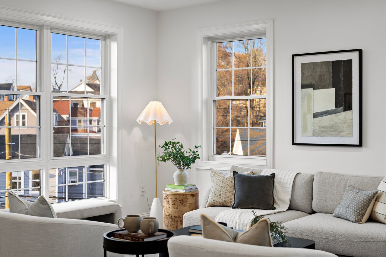 Living room with neutral sofas, large divided-light windows, and autumn trees visible outside.