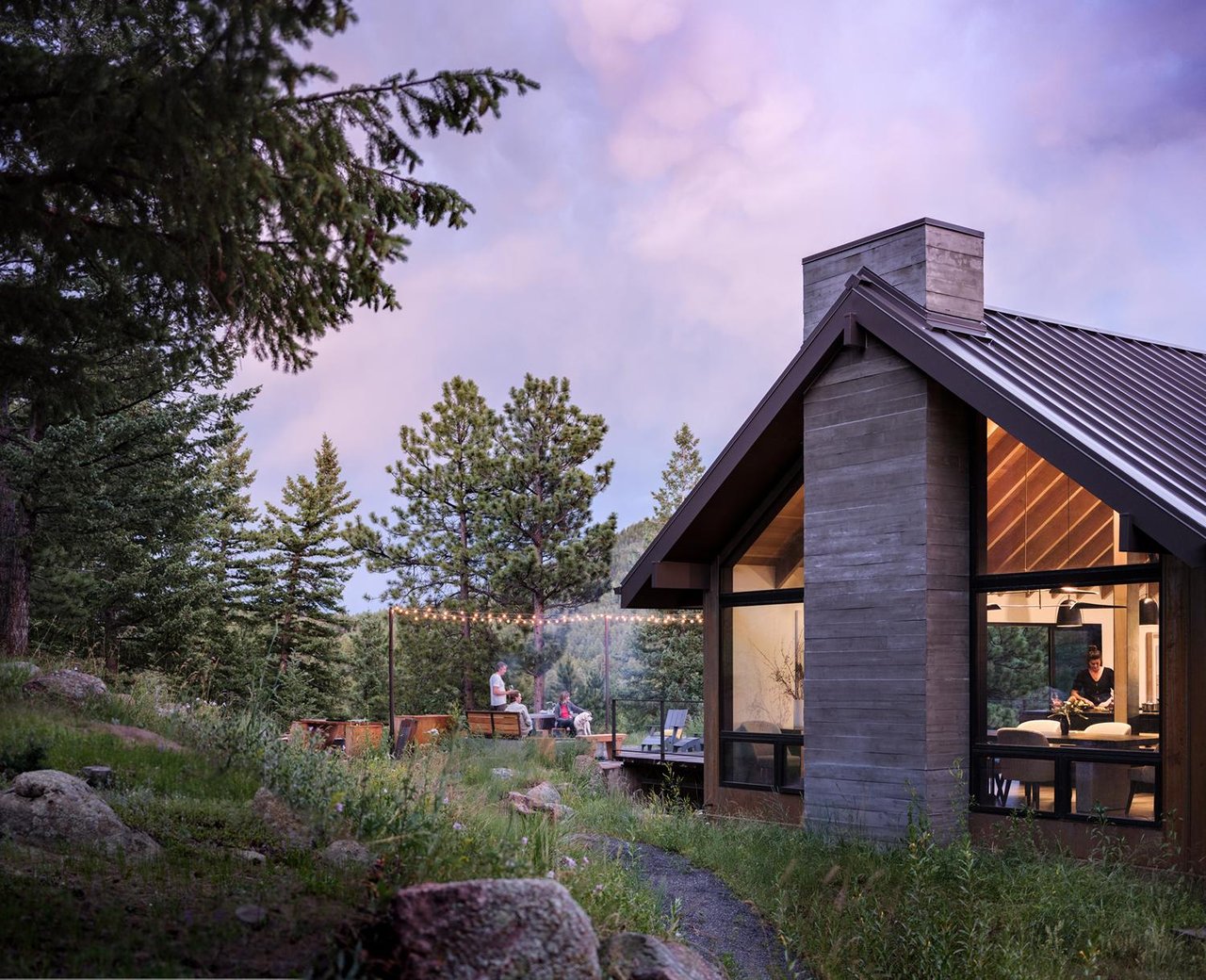 Boulder mountain home at dusk with string lights, outdoor seating, and illuminated interior against purple sky
