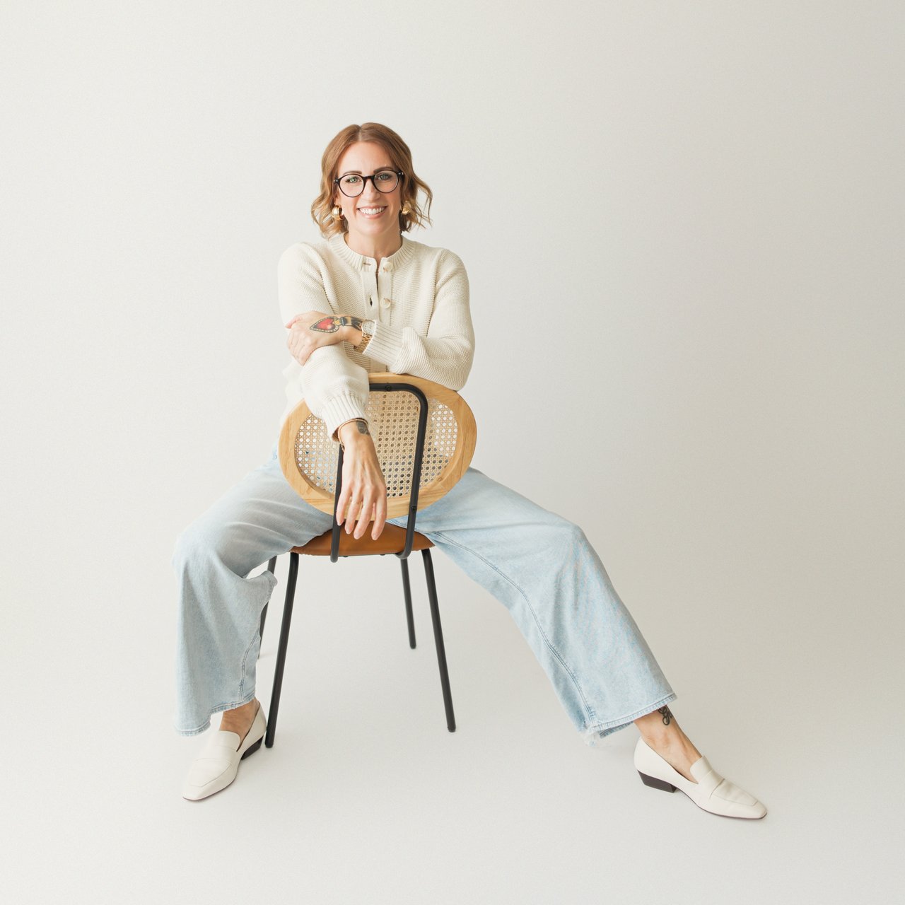 Smiling woman with glasses seated backward on a cane-back chair, wearing a cream sweater and pale jeans.