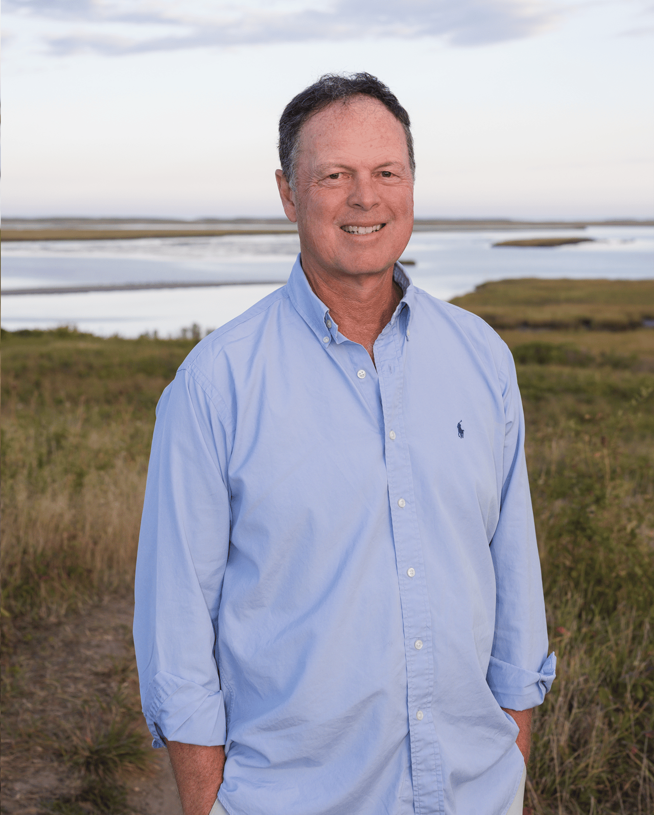 Timothy Waldron standing in a light blue shirt beside a coastal marsh.