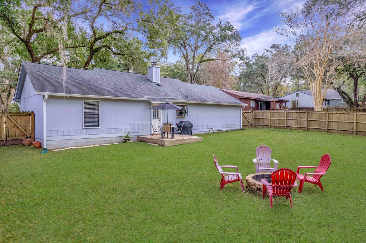 A tranquil backyard with green grass features a cozy fire pit surrounded by colorful Adirondack chairs. A patio has a table, chairs, and an umbrella.