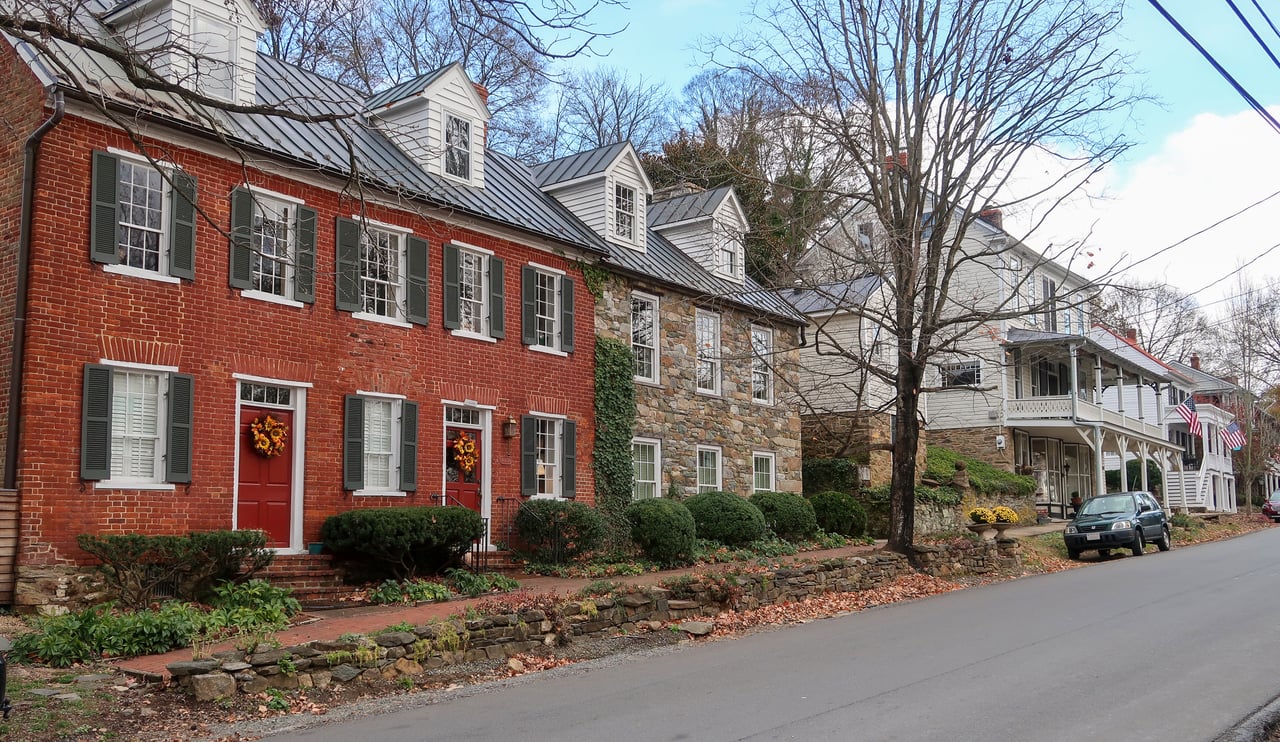 Row of historic brick, stone and clapboard homes with dark shutters and standing-seam metal roofs along a village street in Waterford, Virginia
