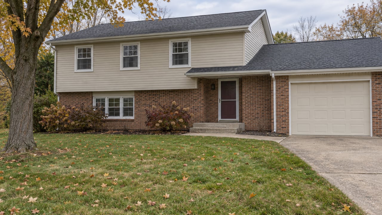 A beige-sided and red-brick split-level home behind a broad autumn lawn, with a mature amber-leaved maple and a concrete driveway.