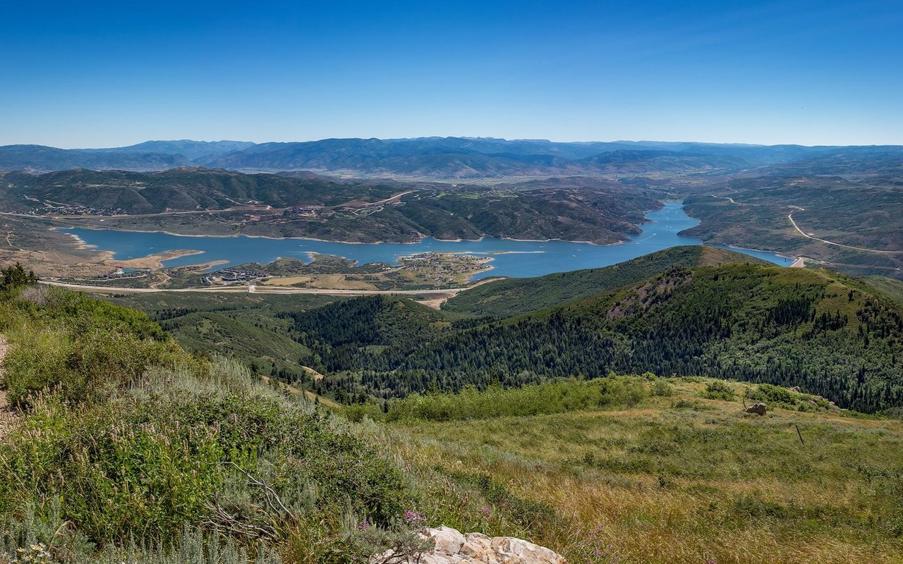Jordanelle Reservoir winds between green hills, viewed from a mountainside with distant ranges beyond.