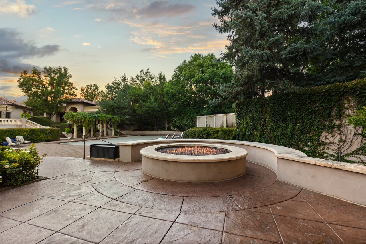 Stone patio with a circular fire pit overlooking a swimming pool and landscaped gardens.