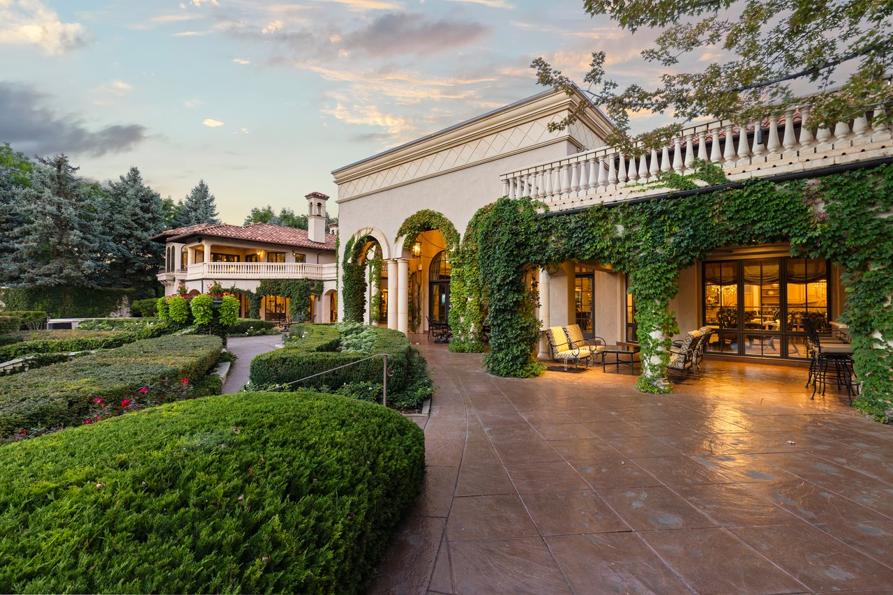 Estate grounds with clipped hedges and flower beds beside an ivy-covered stone patio.