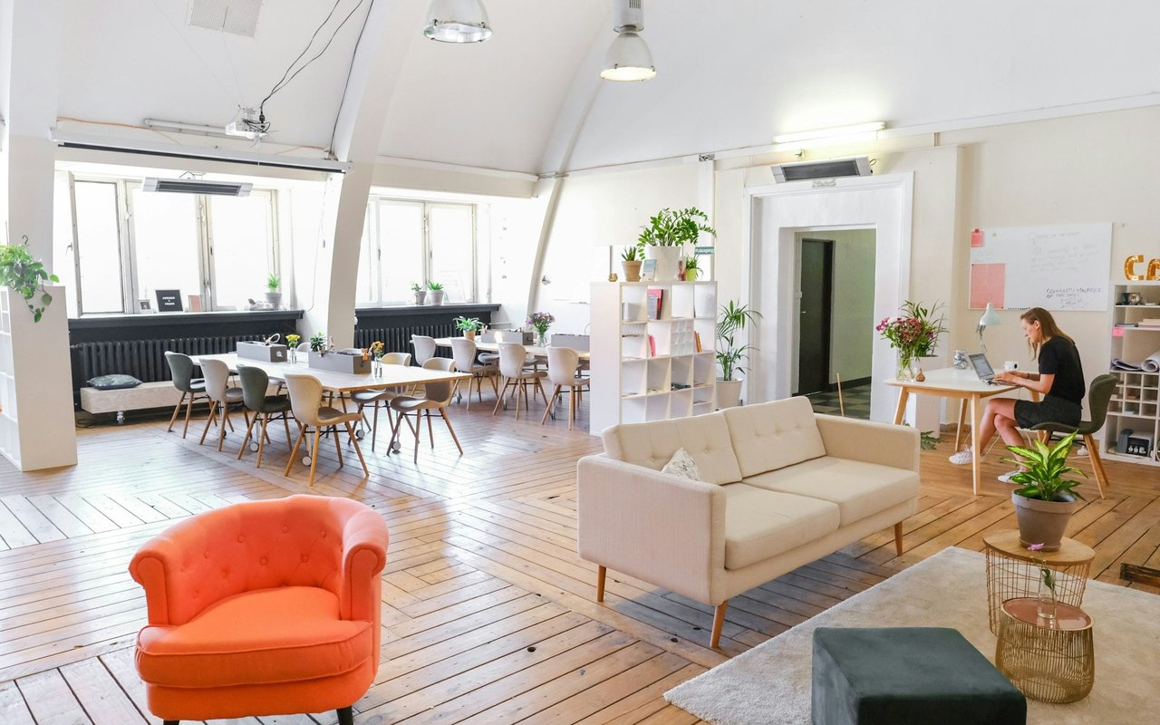 Furnished coworking office with communal tables, lounge seating, and a person working at a desk.