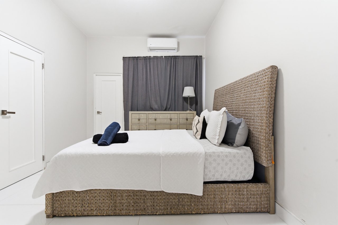 Furnished bedroom with a woven rattan bed, white bedding, gray curtains, and wall-mounted air conditioner.