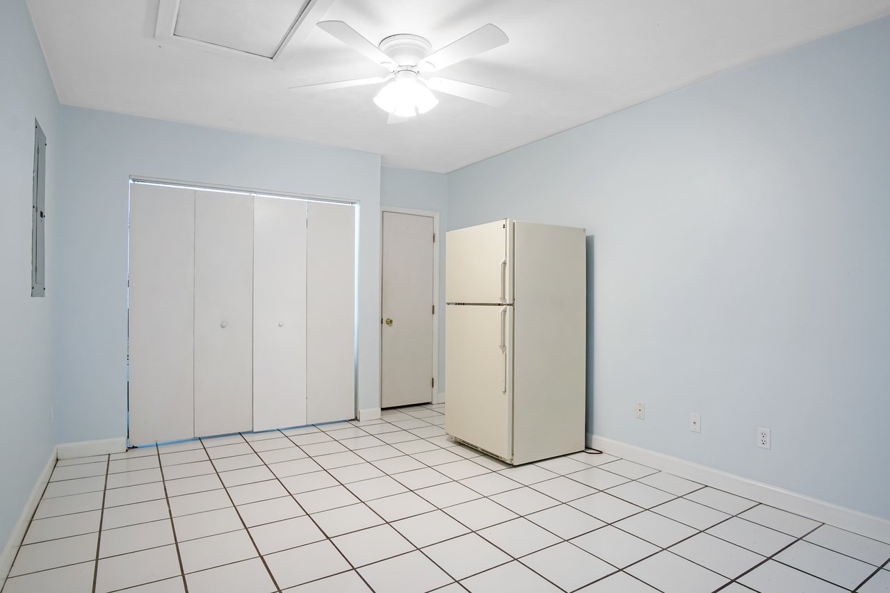 Empty room with a white refrigerator on the right, light blue walls, ceiling fan, and white tiled floor. White closet doors and a closed door are visible.