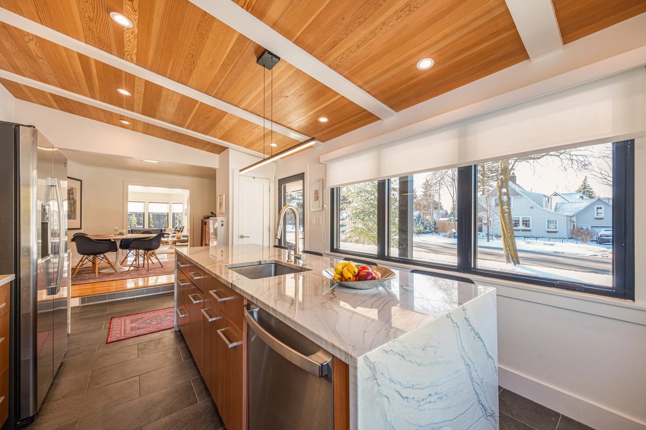 Renovated kitchen with a marble island, wood-paneled ceiling, and large windows overlooking a snowy street.