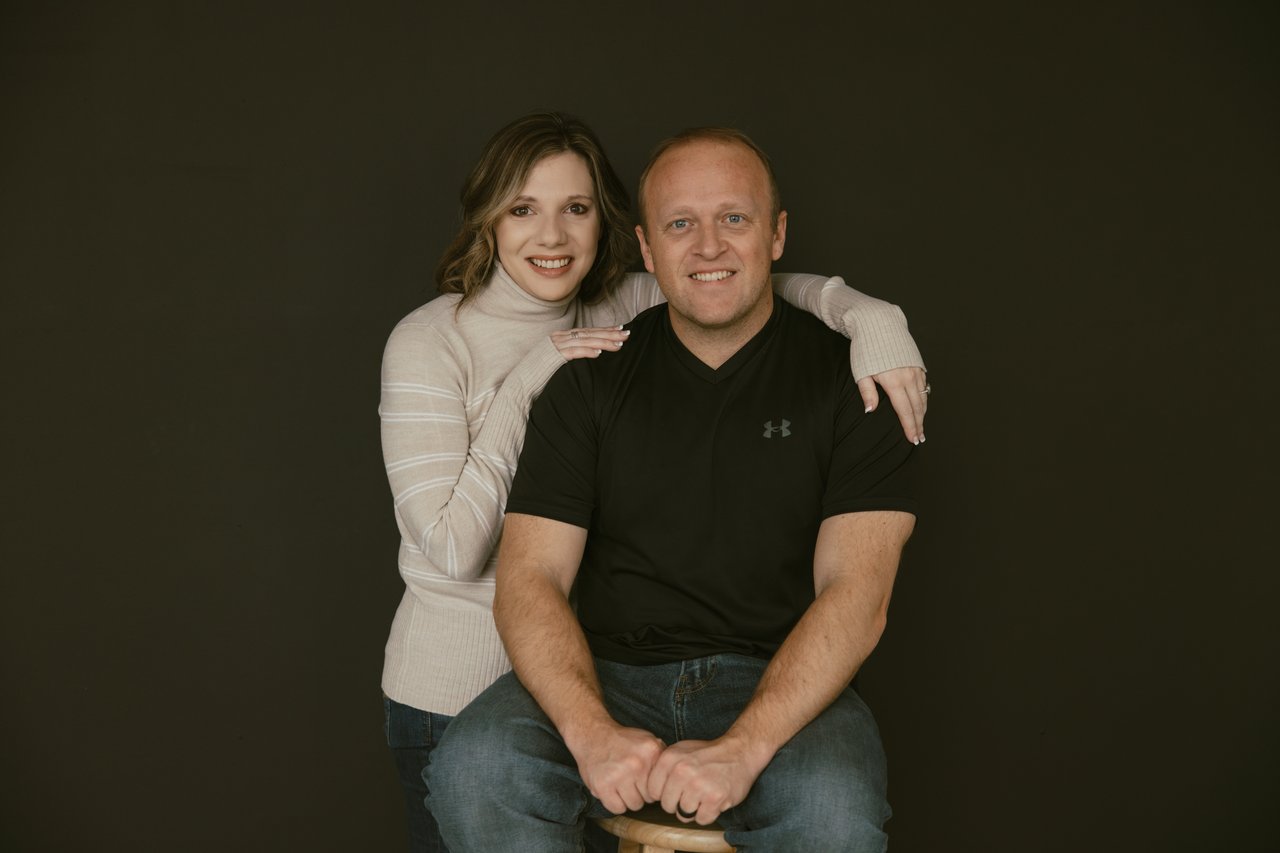 Smiling woman standing behind a seated man in a black shirt against a dark backdrop.