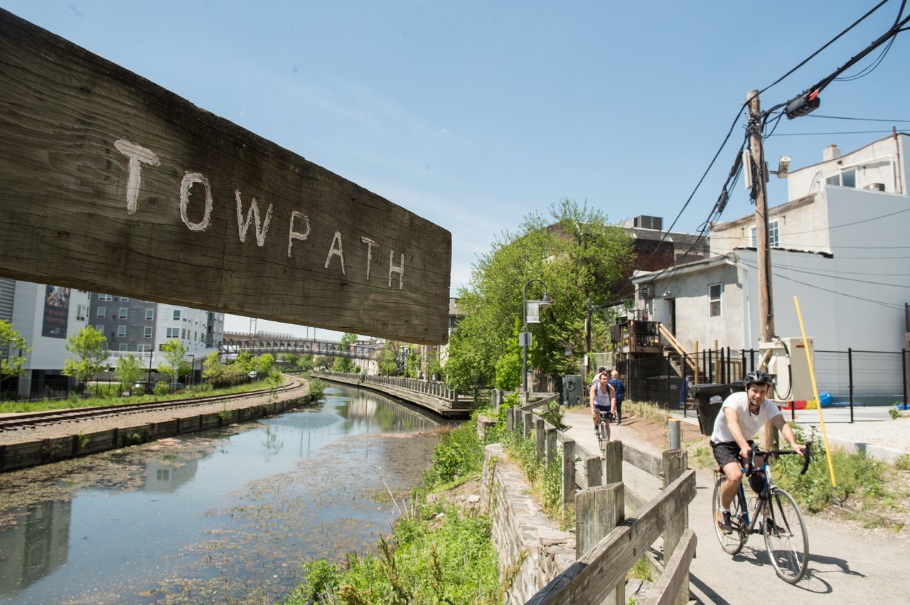 Cyclists ride beside a canal beneath a large wooden TOWPATH sign.