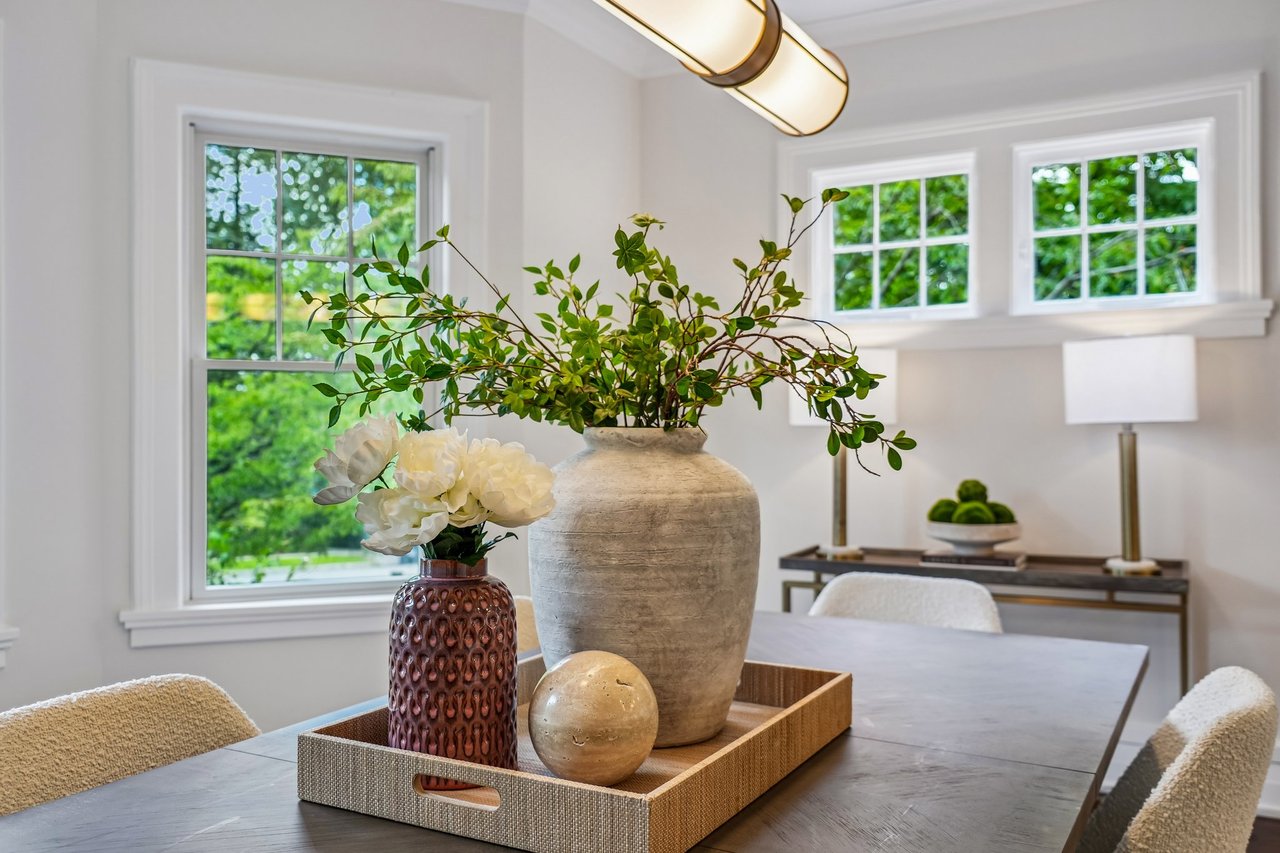 Dining room with a long table, floral centerpiece, and windows overlooking greenery.