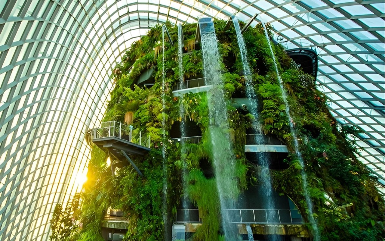 Indoor greenhouse with a plant-covered wall, cascading waterfalls, and elevated walkways beneath a glass roof.