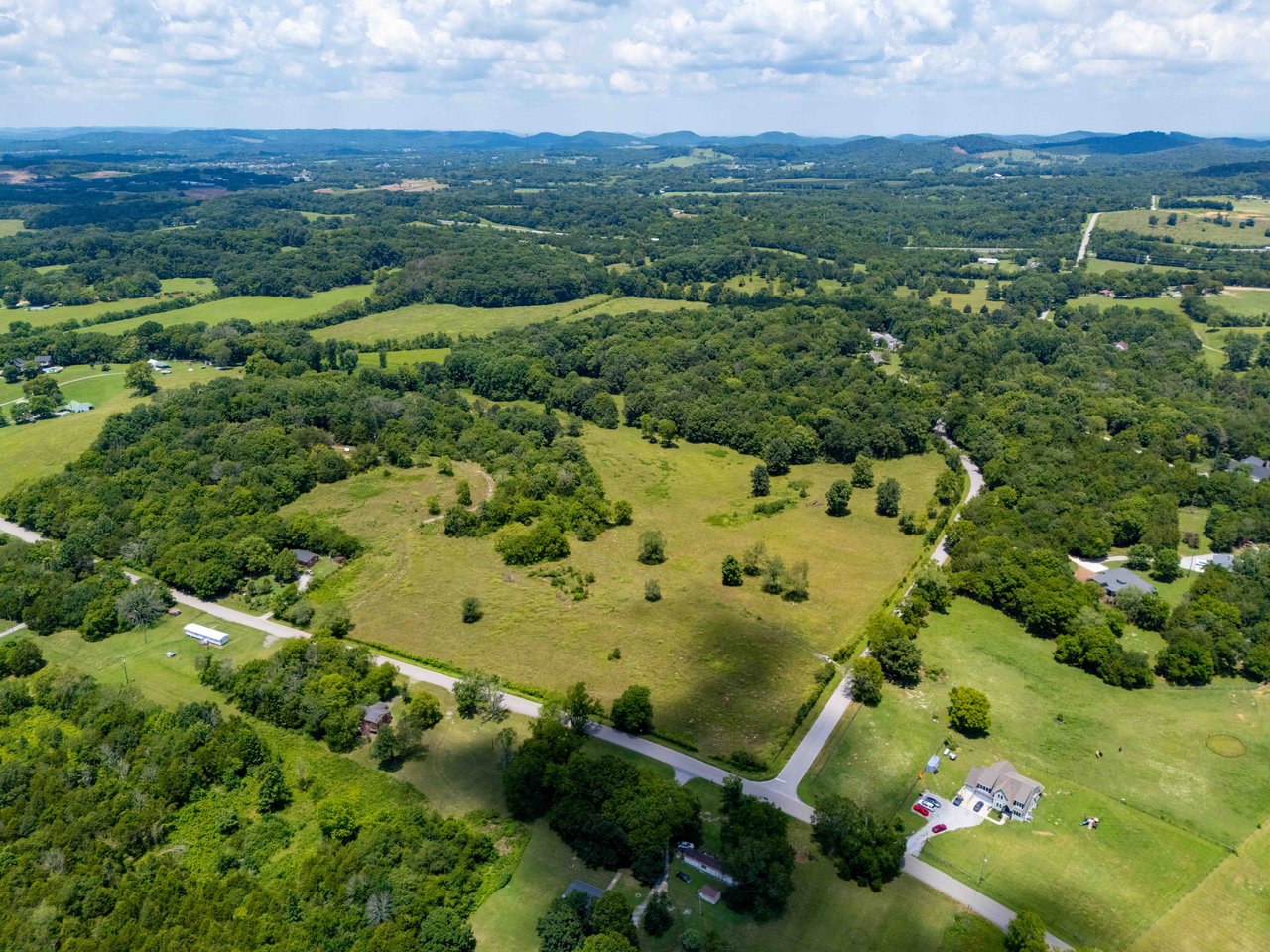 Aerial view of rolling homesite land with open fields, wooded areas, and winding roads.