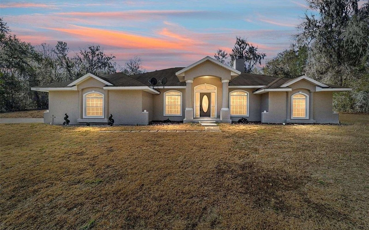 Single-story beige house with illuminated windows, a broad lawn, and a pink-and-blue dusk sky.