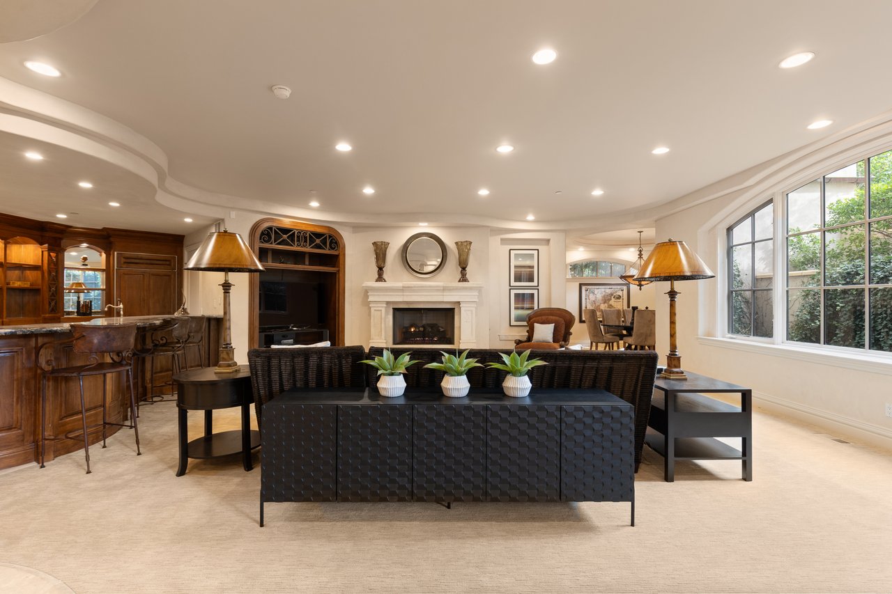 Furnished living room with a white fireplace, dark woven seating, and large windows.