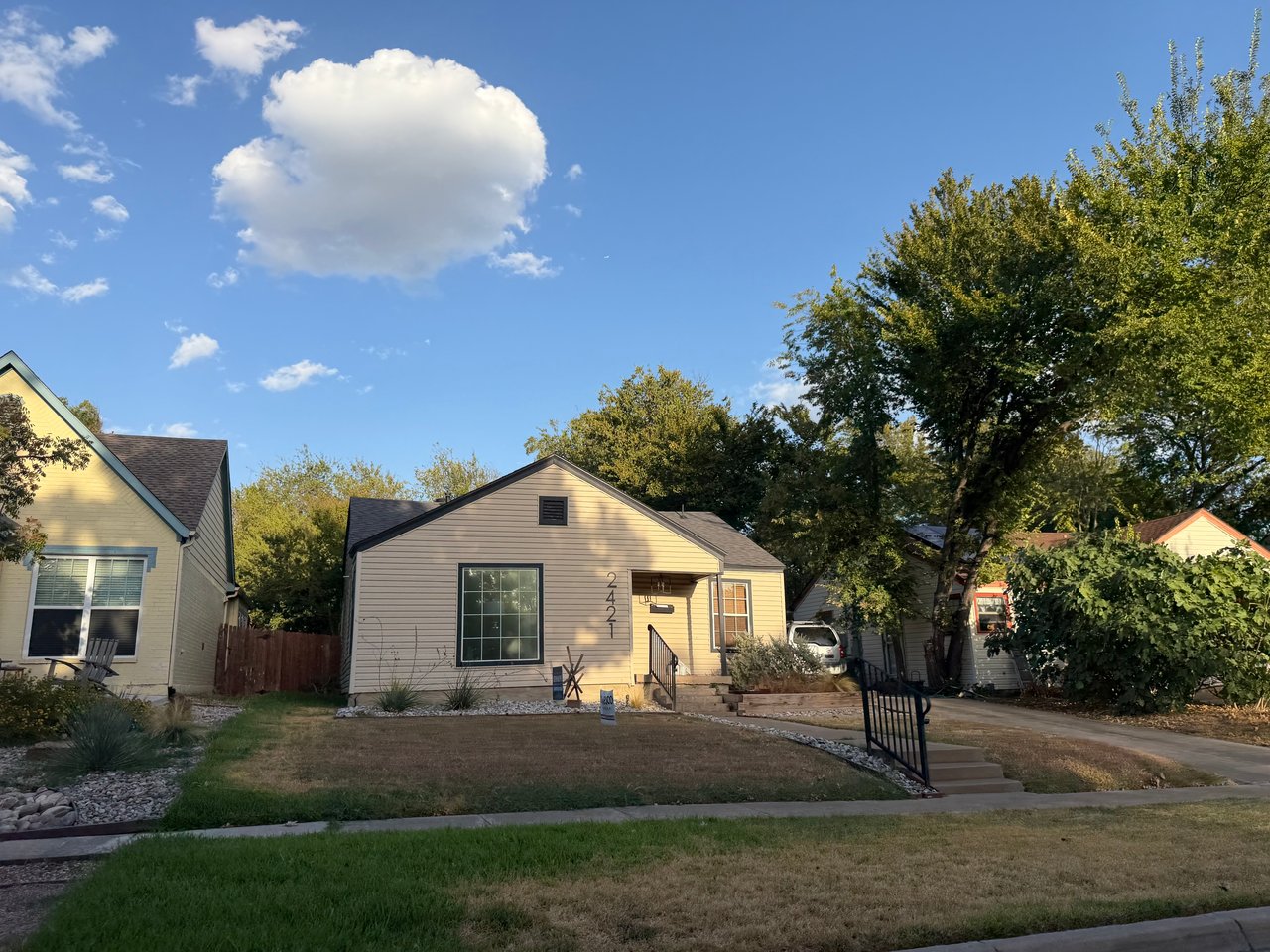 Beige single-story house with a front lawn, driveway, and mature trees; house number 2421.