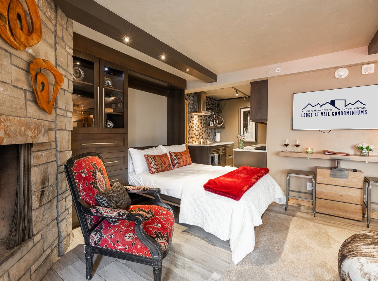 Furnished condominium bedroom with a stone fireplace, patterned red chair, and compact kitchen.
