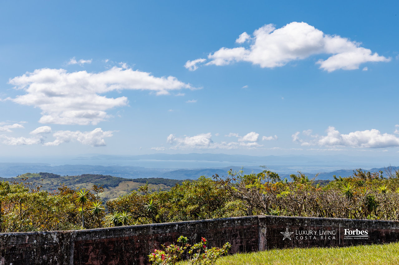 Panoramic mountain view with distant water, foreground shrubs, and a stone wall.