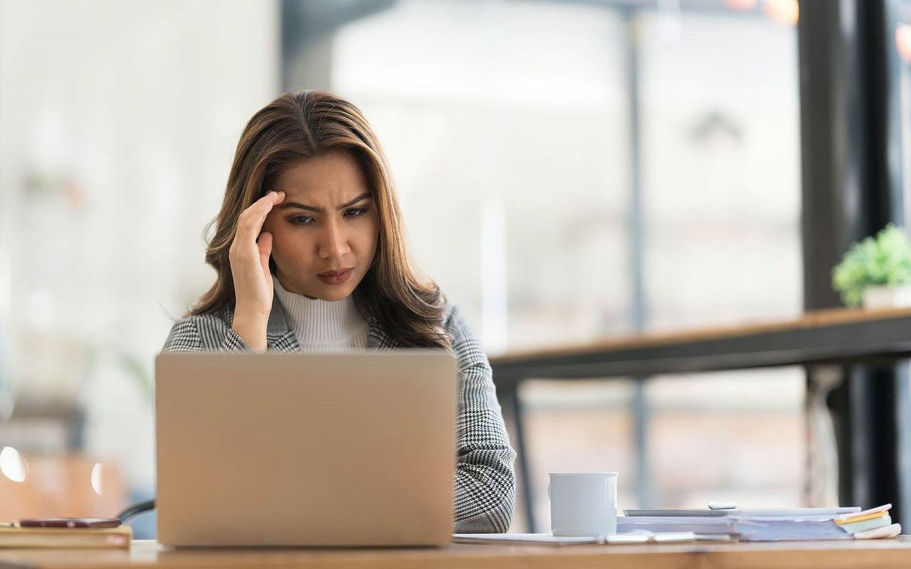 Worried woman touching her temple as she looks at a laptop, with papers and a mug on the desk.