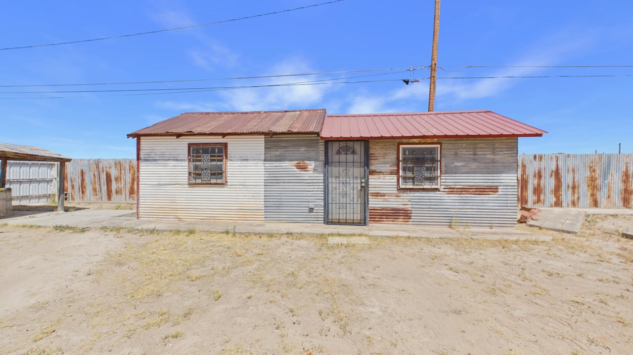 Small metal-sided house with a red roof, barred windows, and a fenced dirt yard.