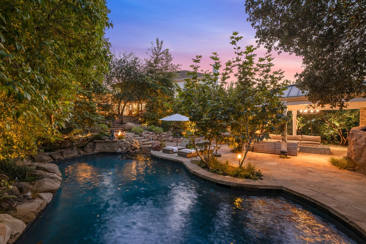 Backyard pool at dusk, bordered by natural stone and a covered outdoor lounge.