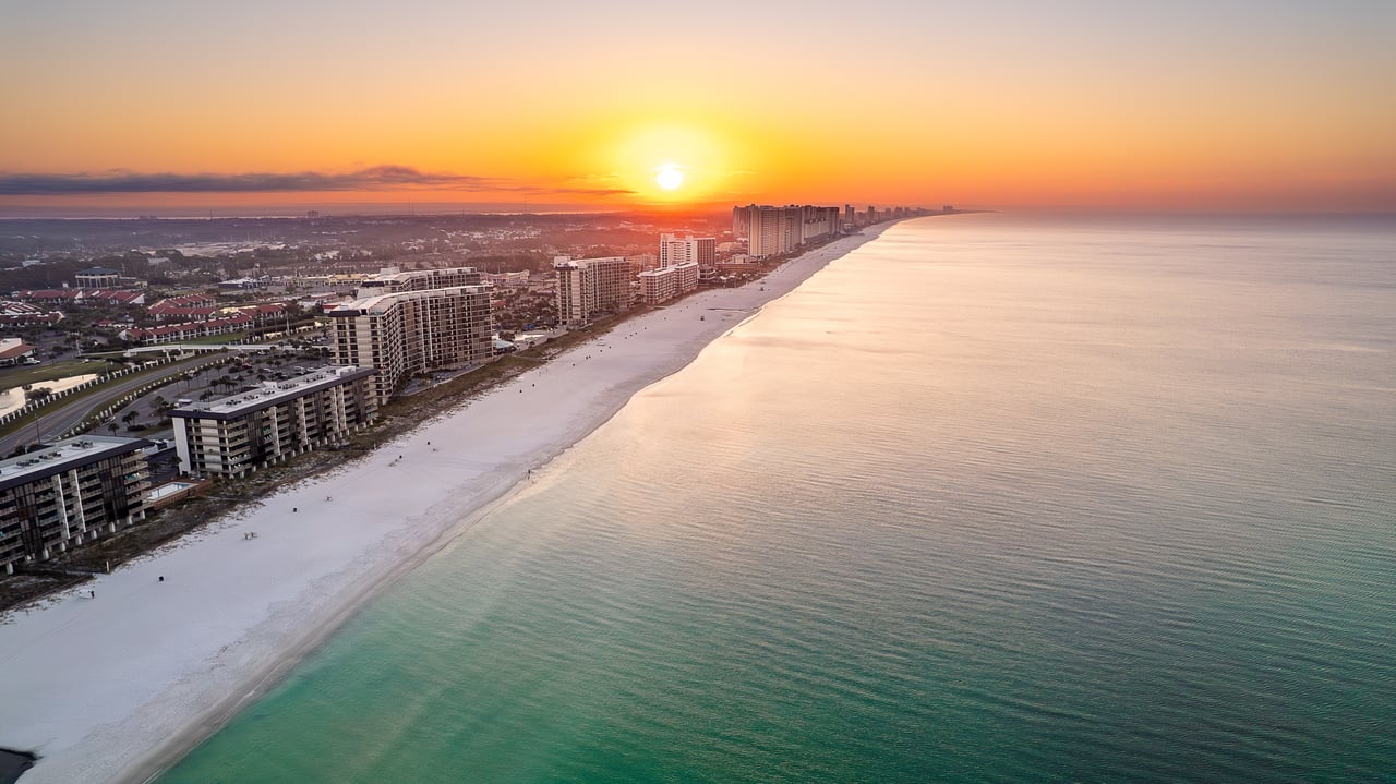 Beachfront condominium towers line a white-sand beach beside calm water at sunset.