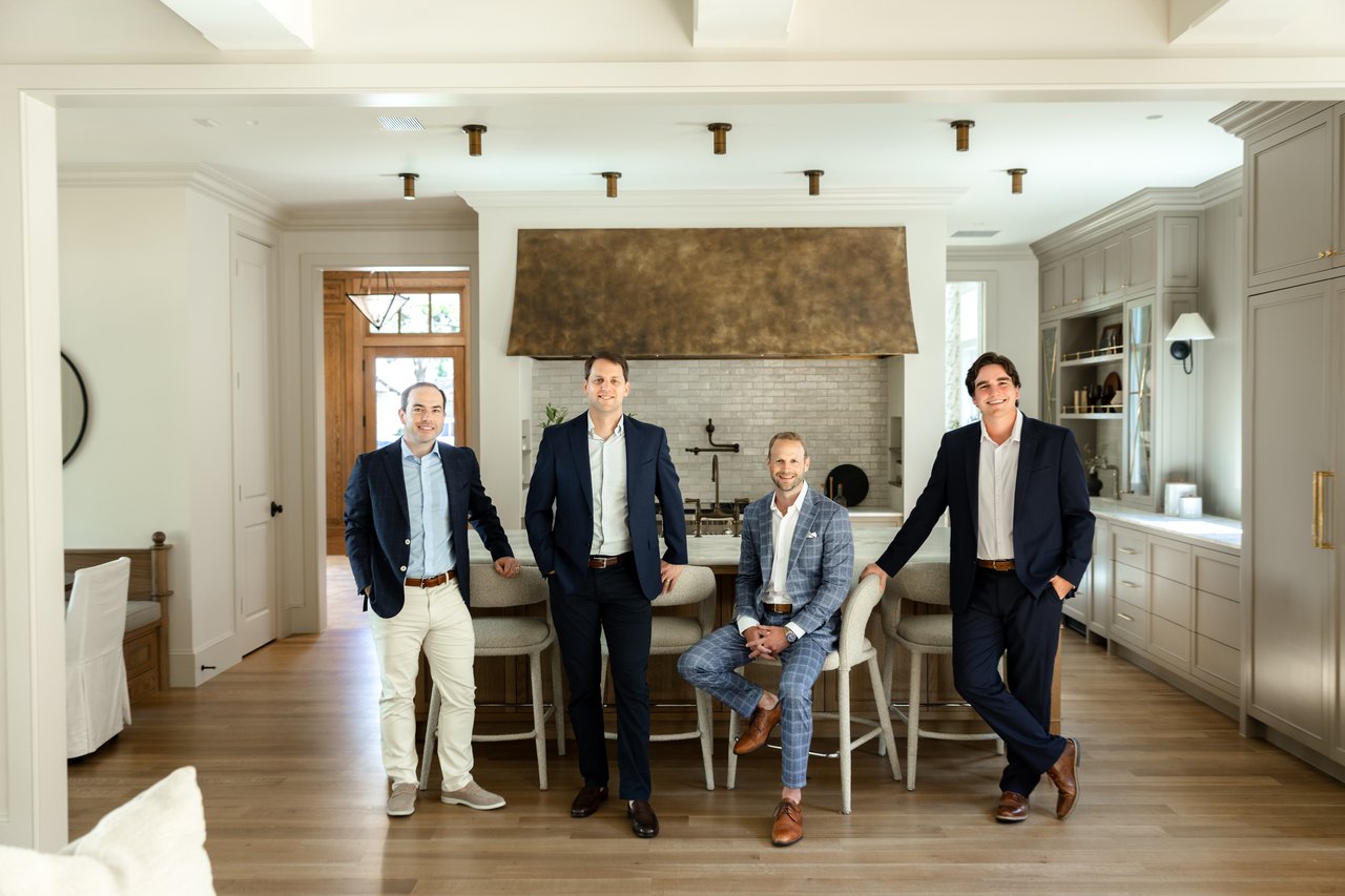 Four real estate team members pose in a bright kitchen, one seated among three standing men.