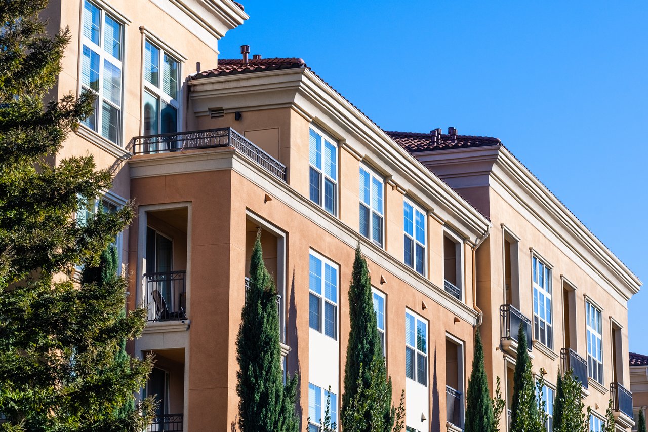 Condominium building with warm stucco walls, tiled roofs, and balconies lined by cypress trees.