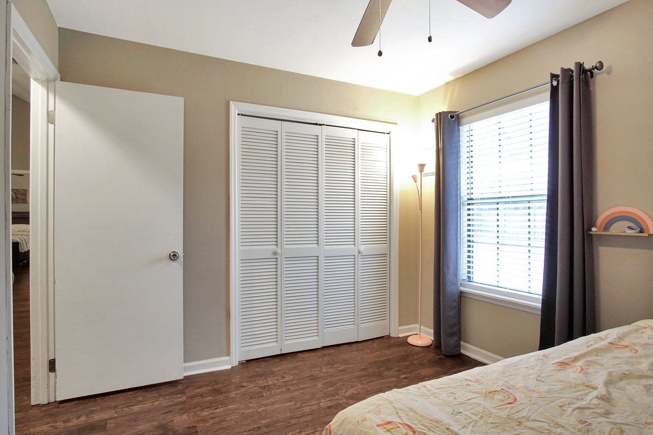 A cozy bedroom features beige walls, a bed with a rainbow-patterned cover, and a white closet with louvered doors. A window with dark curtains lets in natural light.