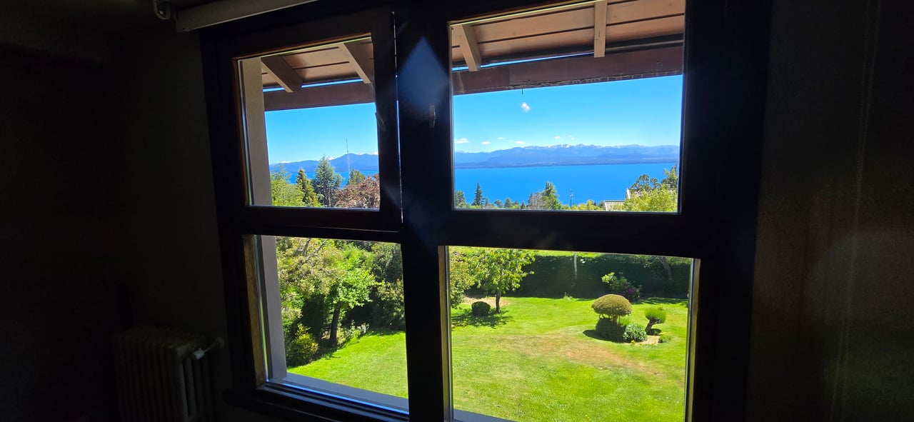 Window view over a landscaped garden toward a blue lake and distant mountains.