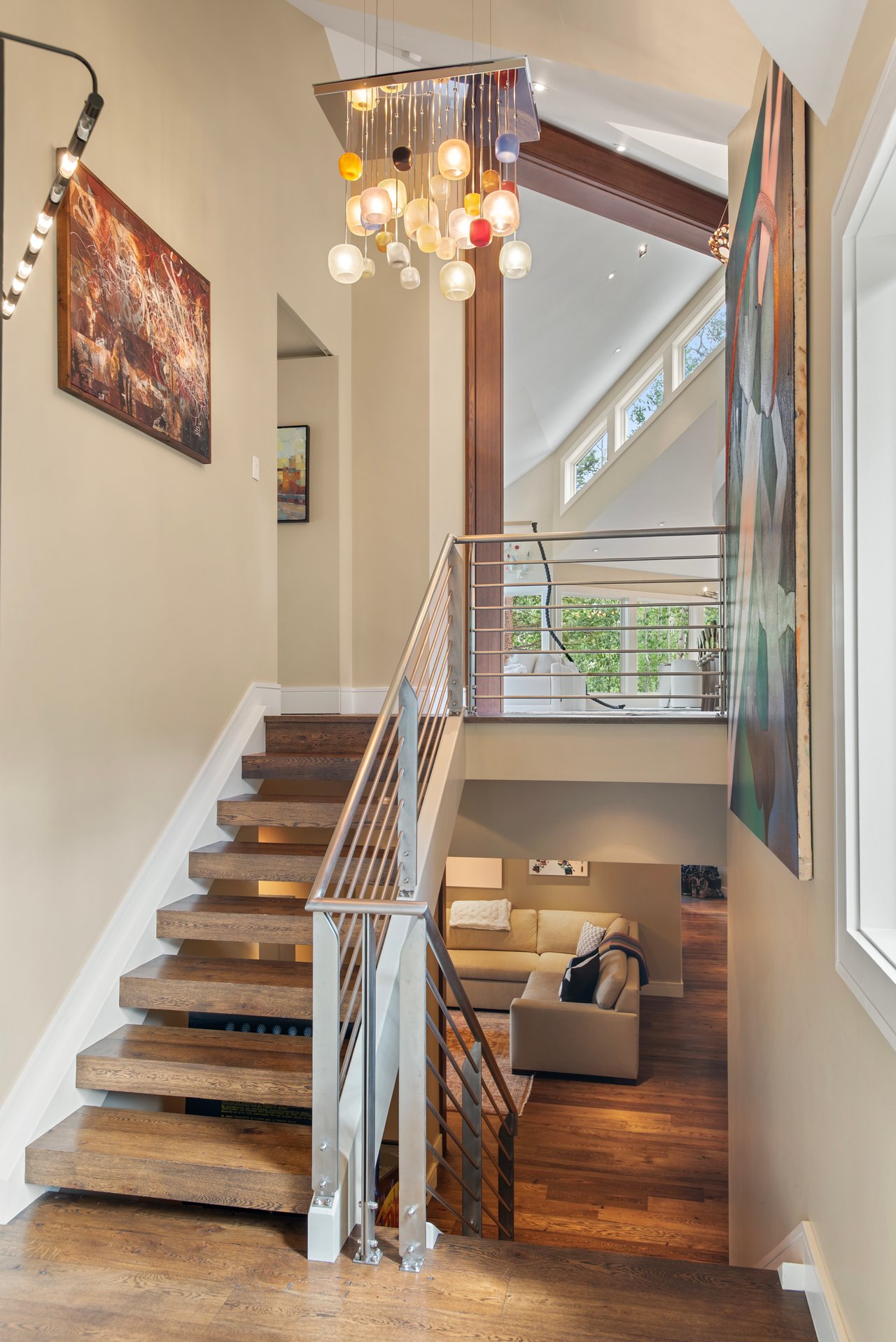 Floating wood staircase with metal railings beneath a colorful chandelier, overlooking a furnished living area.