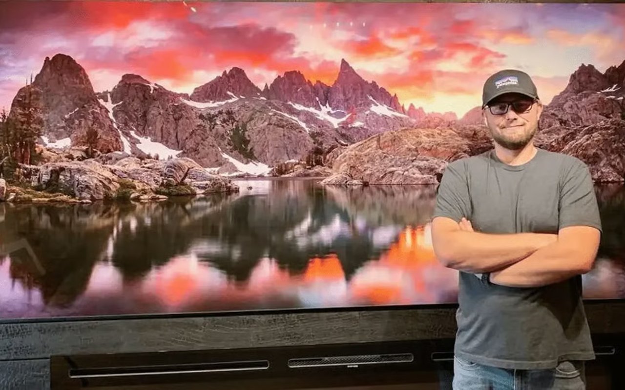 A man with glasses stands arms crossed before a large sunset-lit mountain lake photograph.