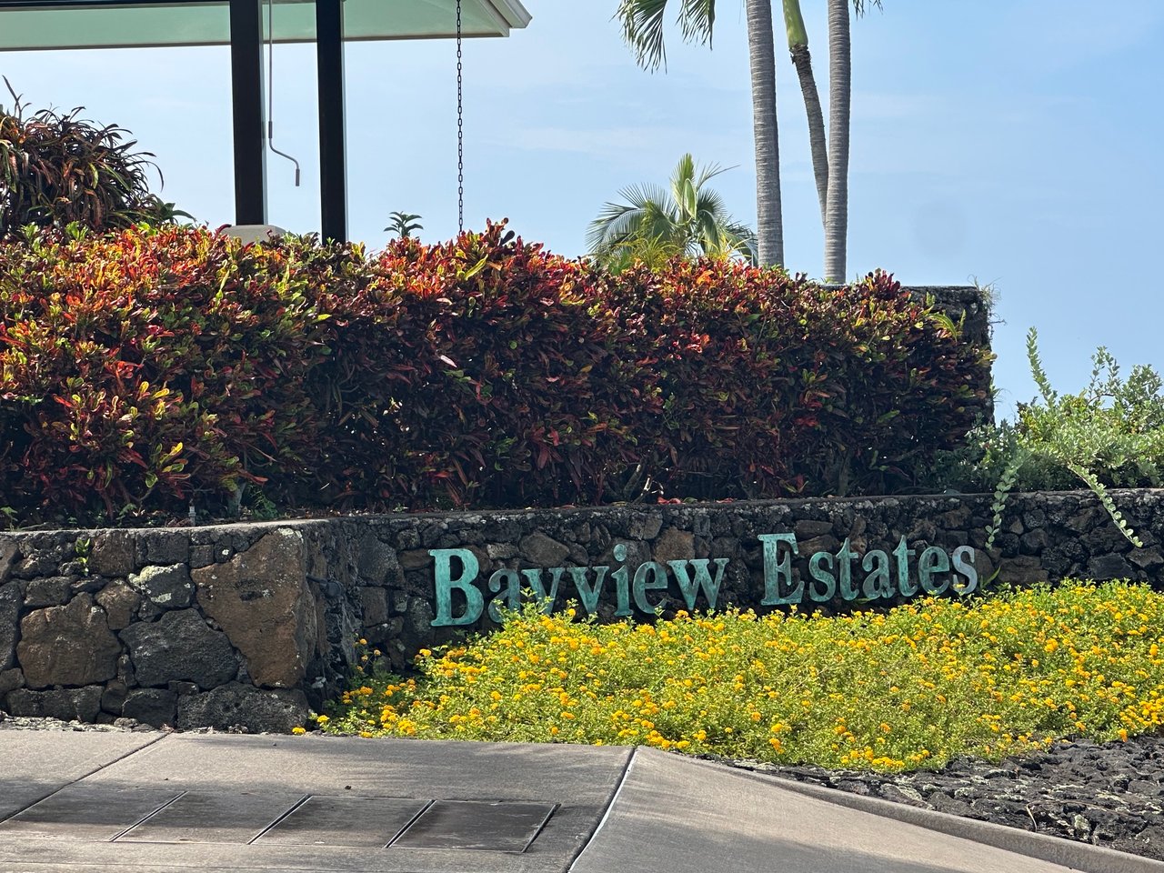 Bayview Estates entrance sign in a lava-rock wall, framed by tropical hedges and yellow flowers.
