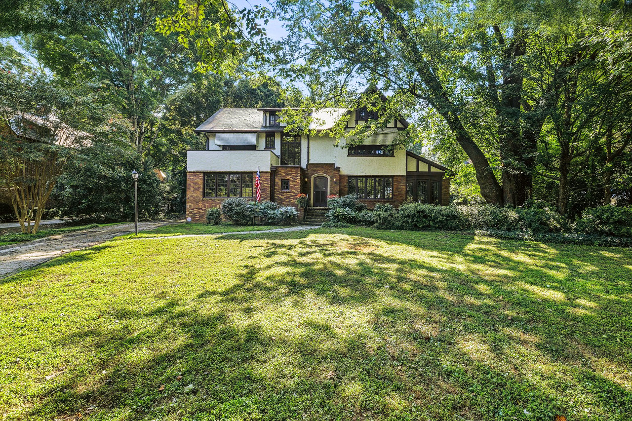 Two-story brick-and-siding house framed by mature trees, with a broad lawn and American flag by the entrance.