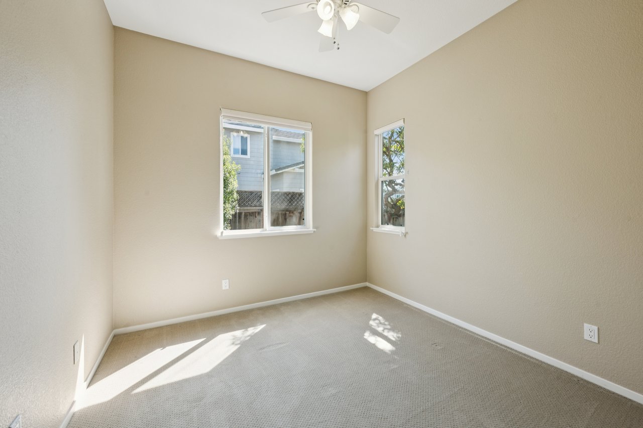 Empty bedroom with beige walls, two windows, and sunlight across the carpet.