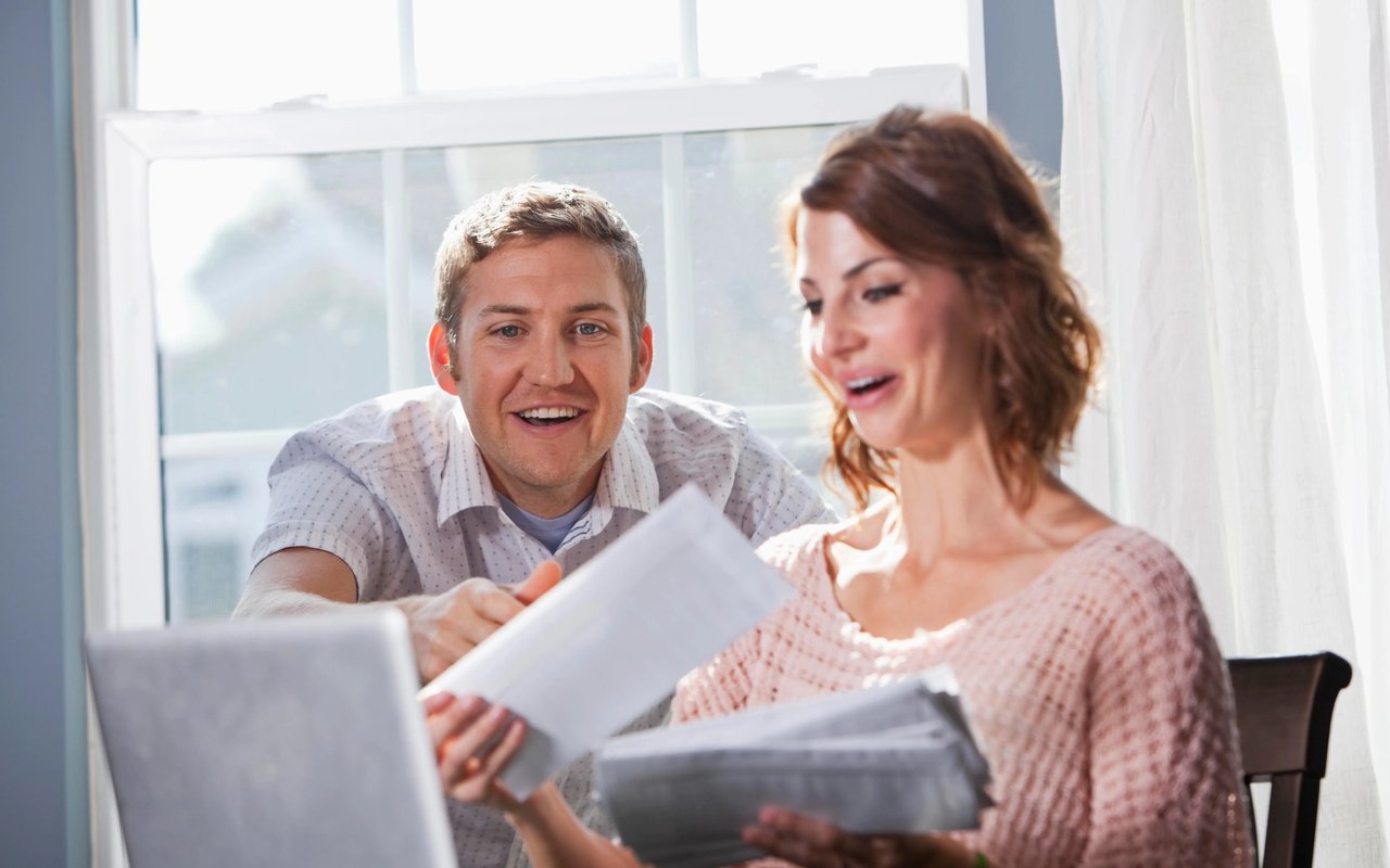 A couple reviews paperwork together beside an open laptop.