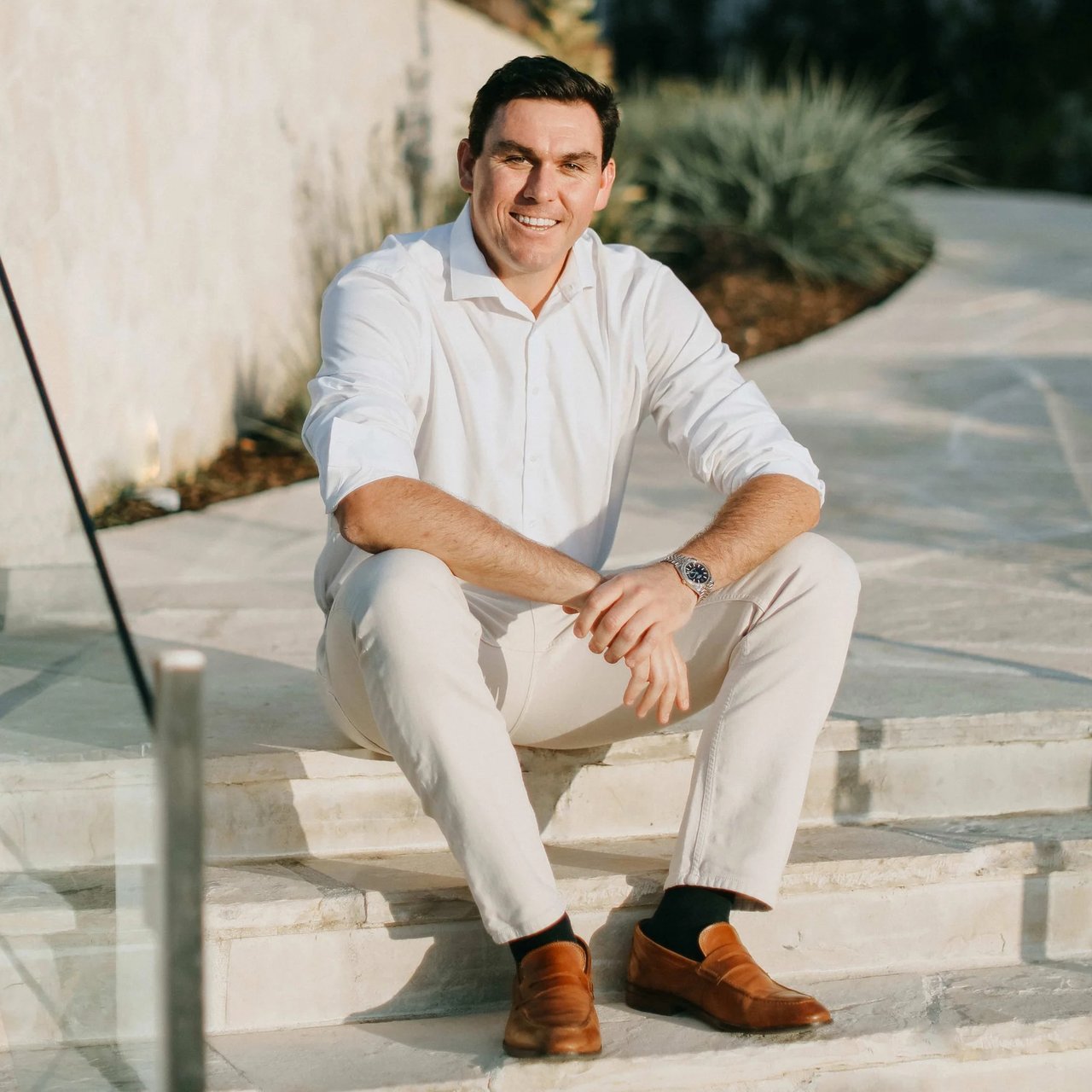 Man in a white shirt seated on sunlit stone steps, wearing brown loafers and a wristwatch.