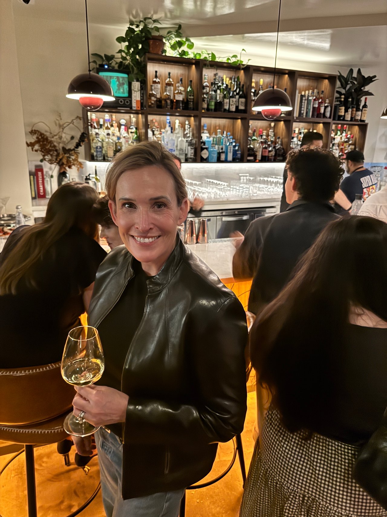 Smiling woman in a black leather jacket holding a wine glass at a crowded bar.