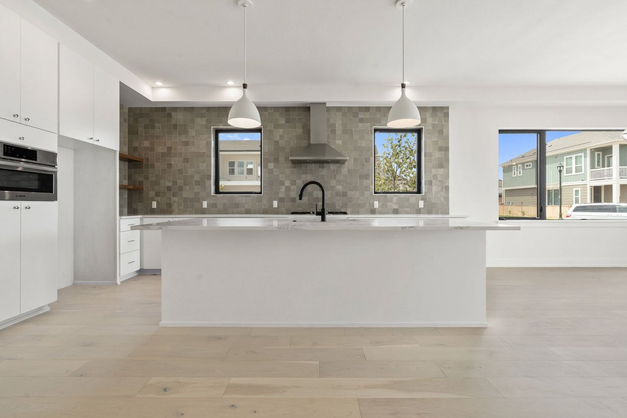 Modern kitchen with a large white island, gray tile backsplash, and windows overlooking neighboring homes.